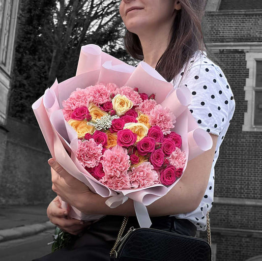 Person holding a bouquet of pink and yellow flowers outdoors.