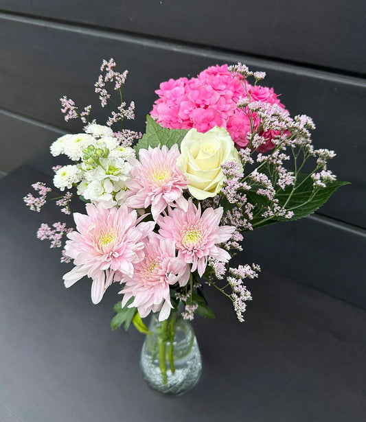 Bouquet of pink, white, and green flowers in a clear vase on a dark surface.